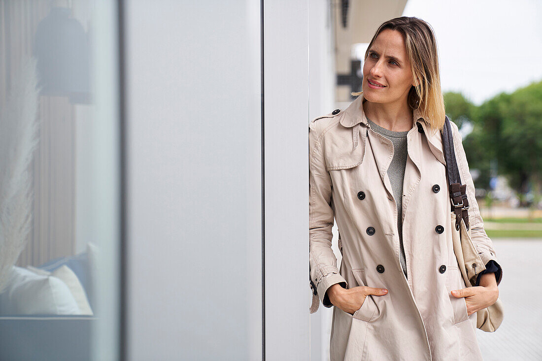 Mid-shot of woman wearing raincoat looking at shop window while leaning on a wall