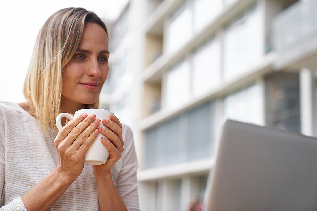 Mittlere Frontalaufnahme einer Frau mit einer Tasse Kaffee in der Hand und Blick auf den Bildschirm eines Laptops