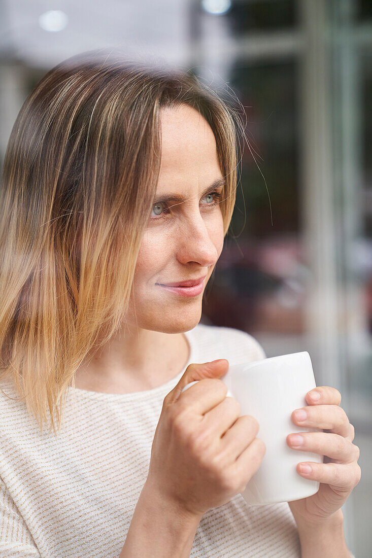 Porträt einer Frau, die einen Kaffeebecher hält und durch ein Fenster schaut