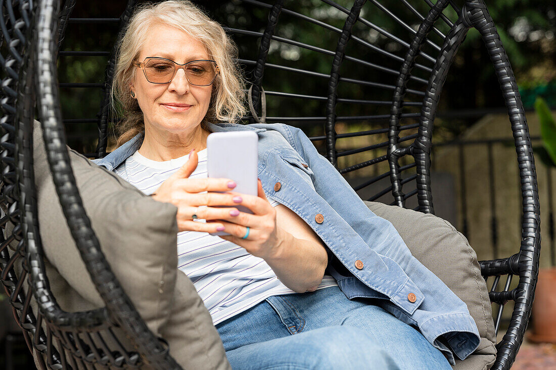 Ältere Frau, die in einem bequemen Gartenstuhl sitzt und ihre Nachrichten auf dem Mobiltelefon überprüft