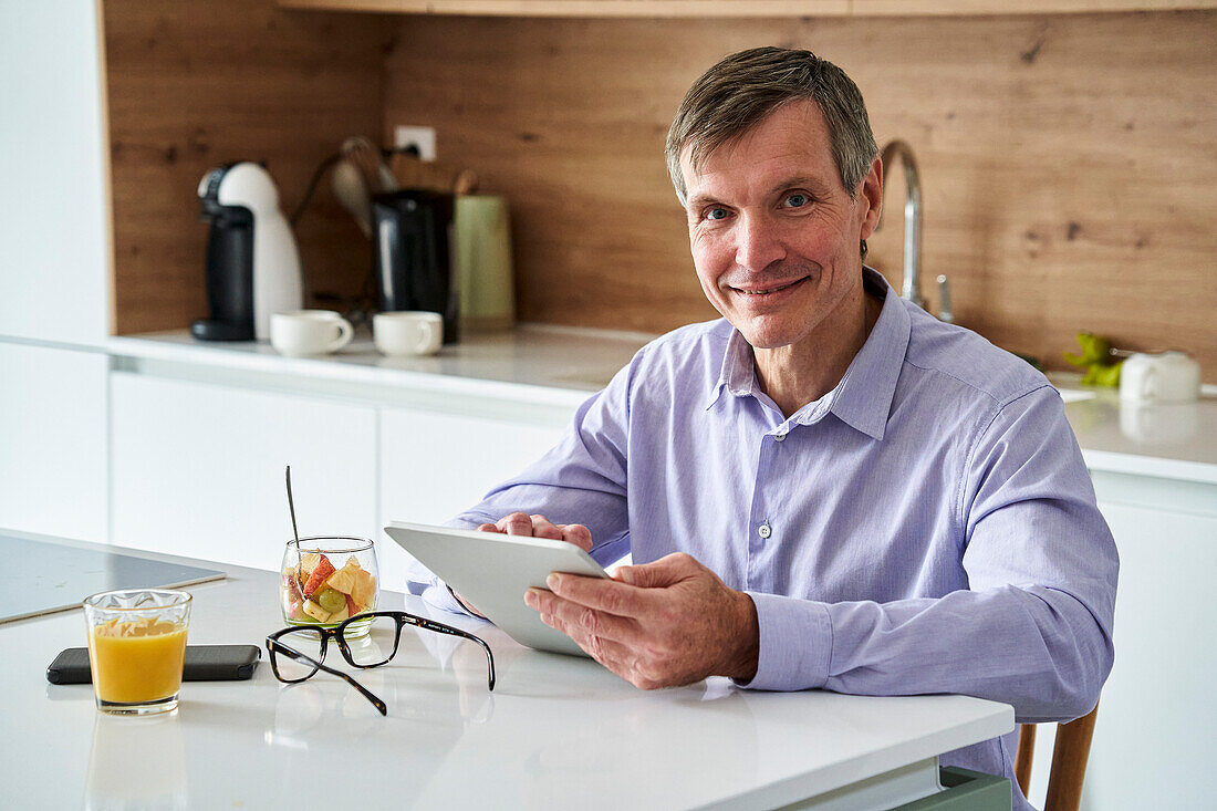 Middle-aged professional man looking at camera and checking messages on digital tablet while having breakfast on kitchen counter at home