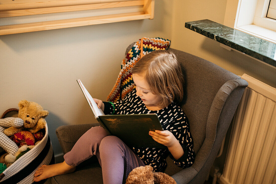 Girl sitting in armchair and reading book at home