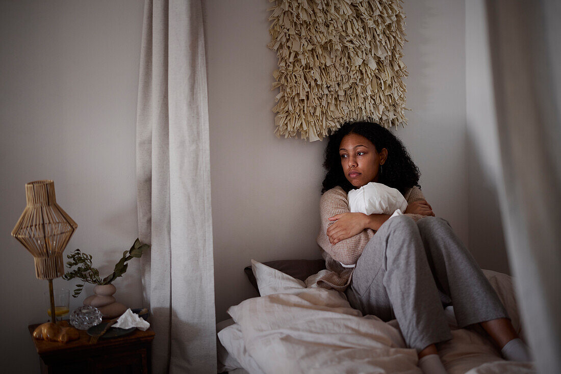 Pensive young woman sitting on bed and hugging pillow