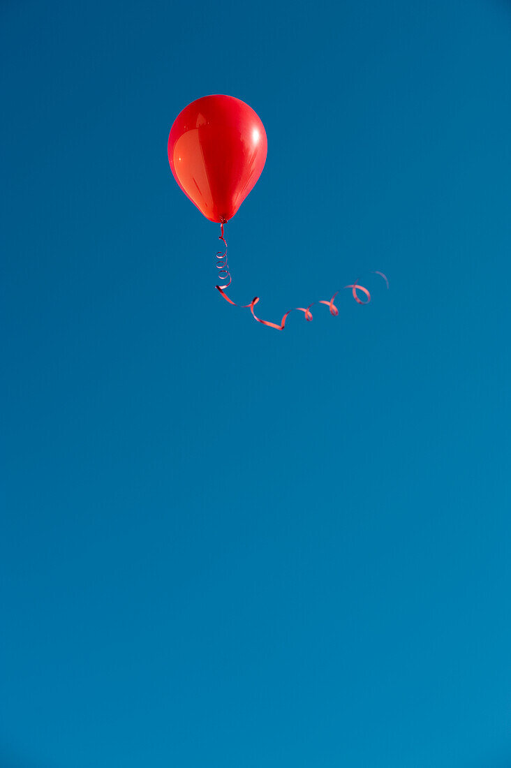 Roter Luftballon mit Schleife fliegt gegen blauen Himmel