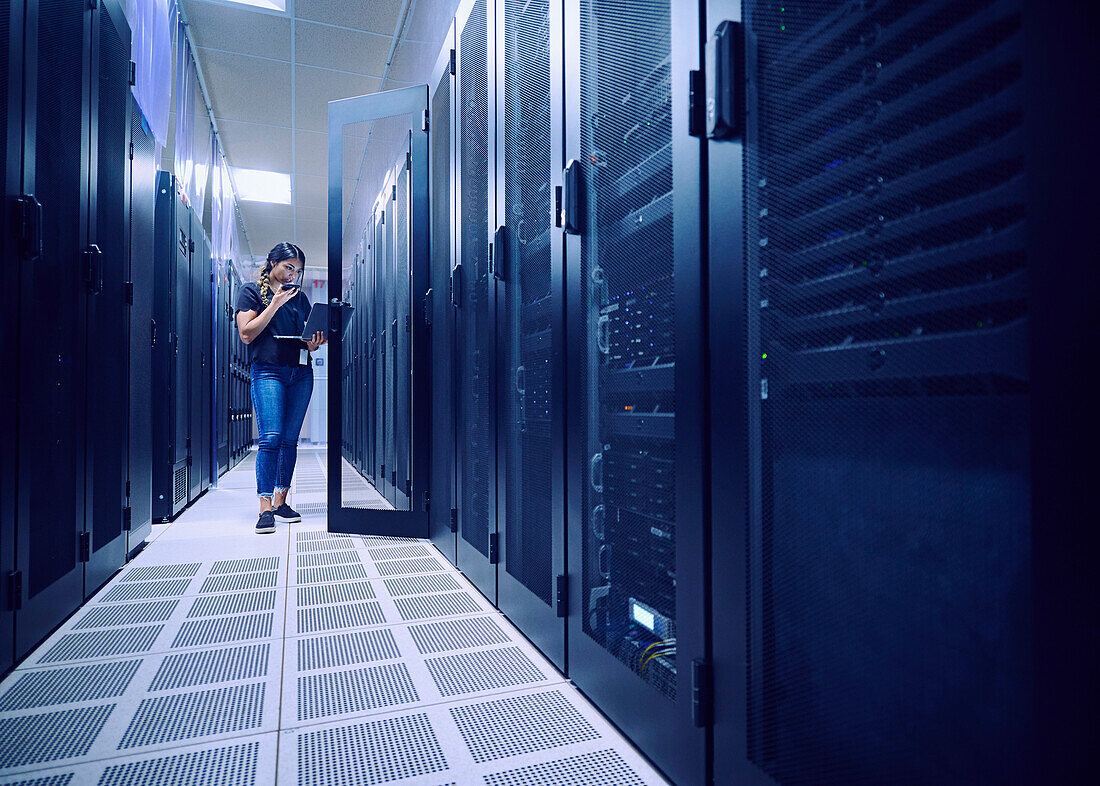 Female technician using laptop and phone in server room