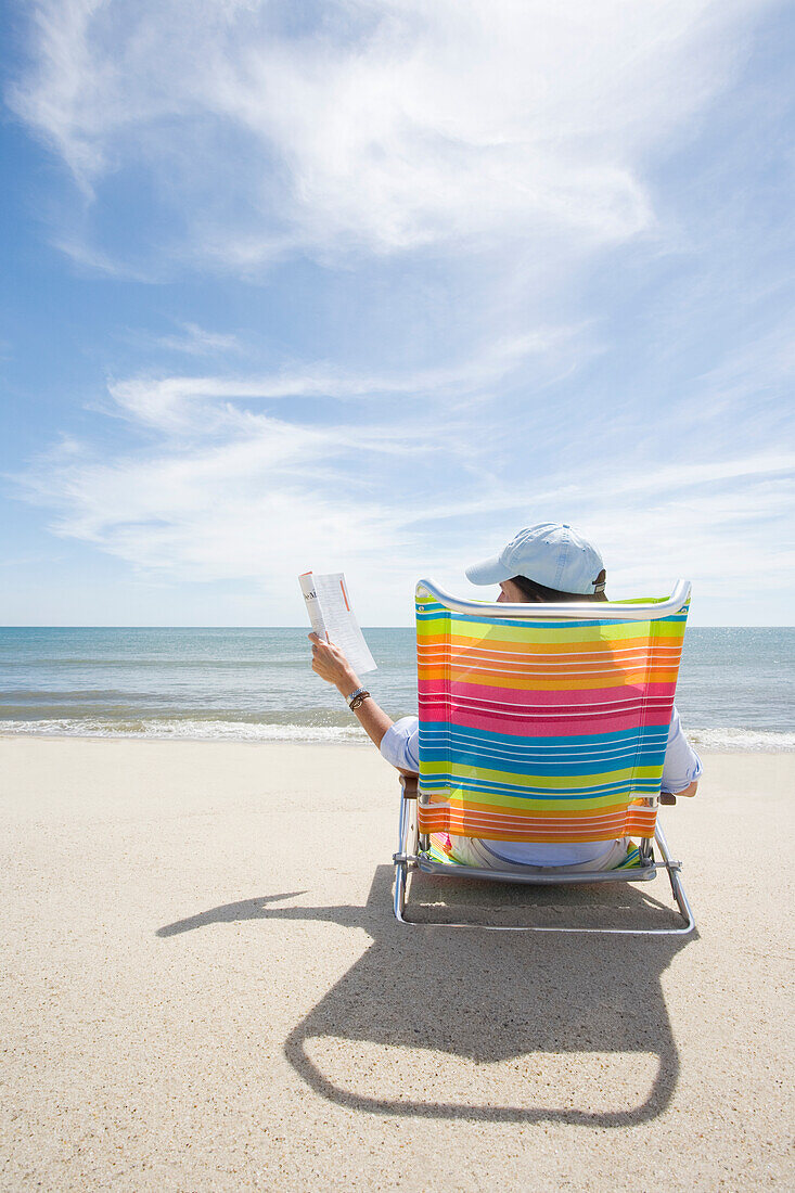 USA, Massachusetts, Cape Cod, Nantucket Island, Rückansicht einer Frau in einem bunten Strandkorb, die am Strand eine Zeitschrift liest