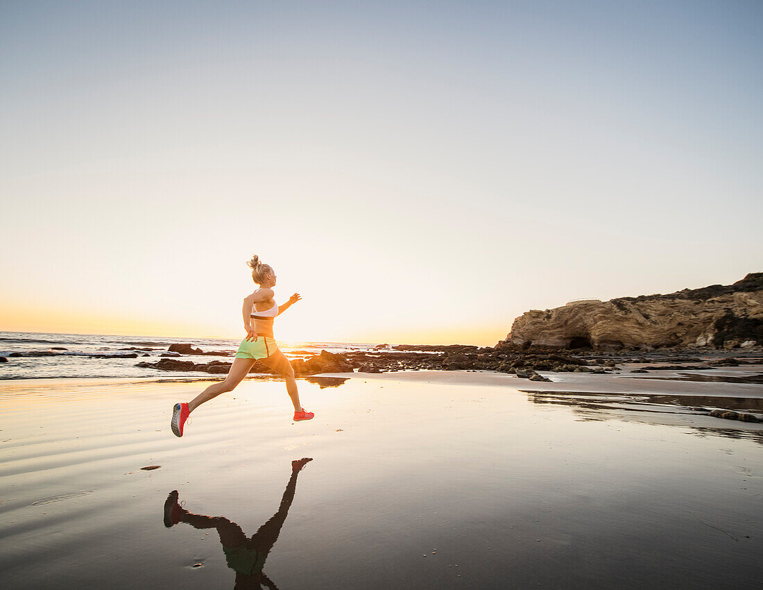 Vereinigte Staaten, Kalifornien, Laguna Beach, Rückansicht einer Sportlerin, die bei Sonnenuntergang am Strand läuft