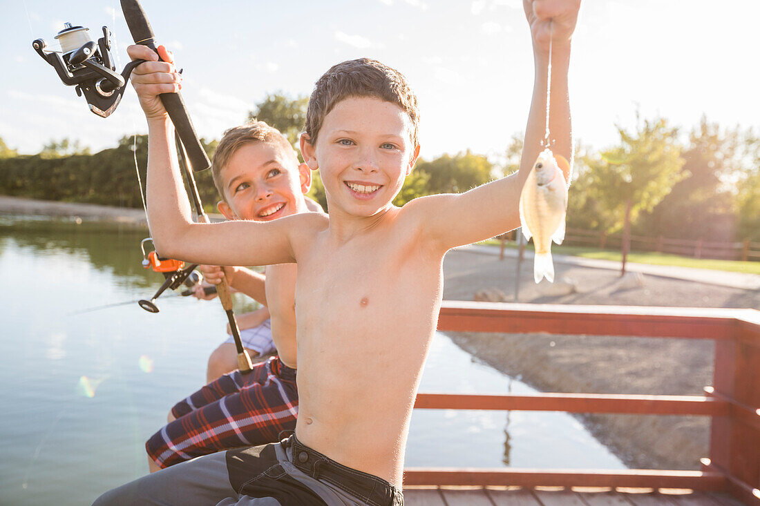 Portrait of smiling shirtless boy (8-9) holding fish and fishing rod by lake