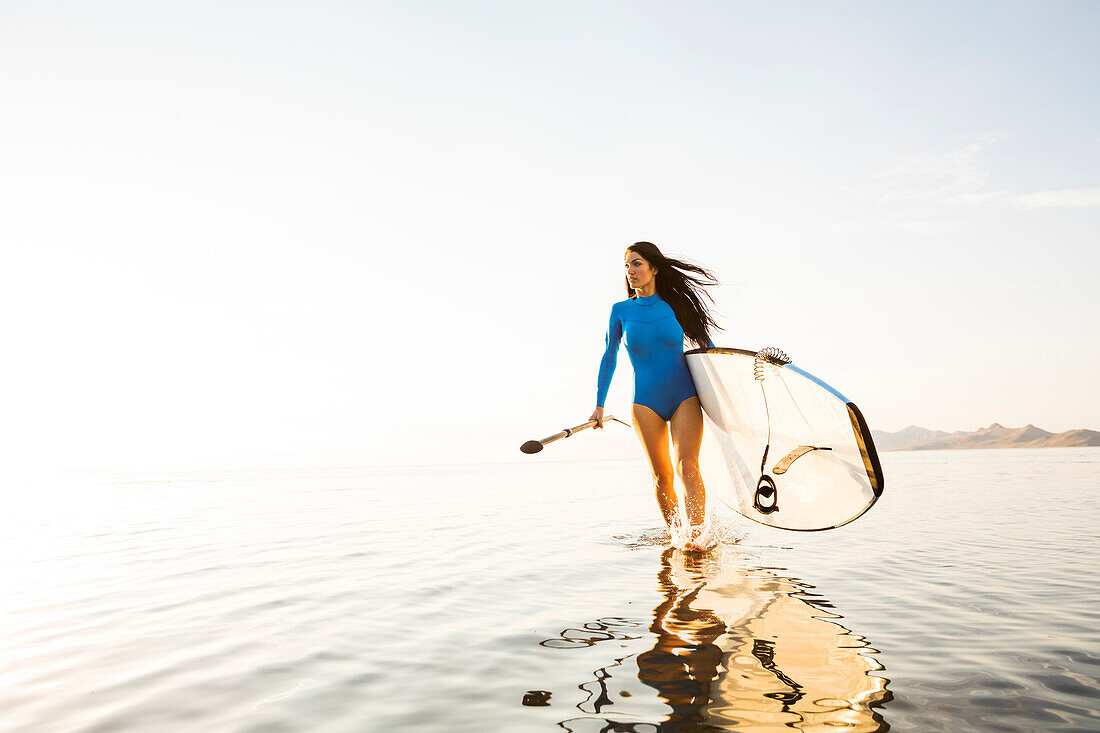 Woman in blue swimsuit carrying paddleboard in lake at sunset