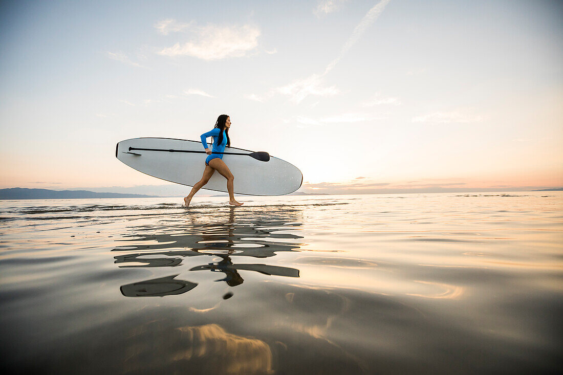 Frau läuft am Strand mit Paddleboard bei Sonnenuntergang 
