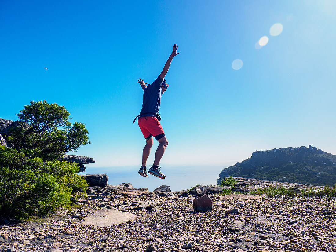 Südafrika, Westkap, Kapstadt, Mann springt mit erhobenen Armen auf eine Klippe