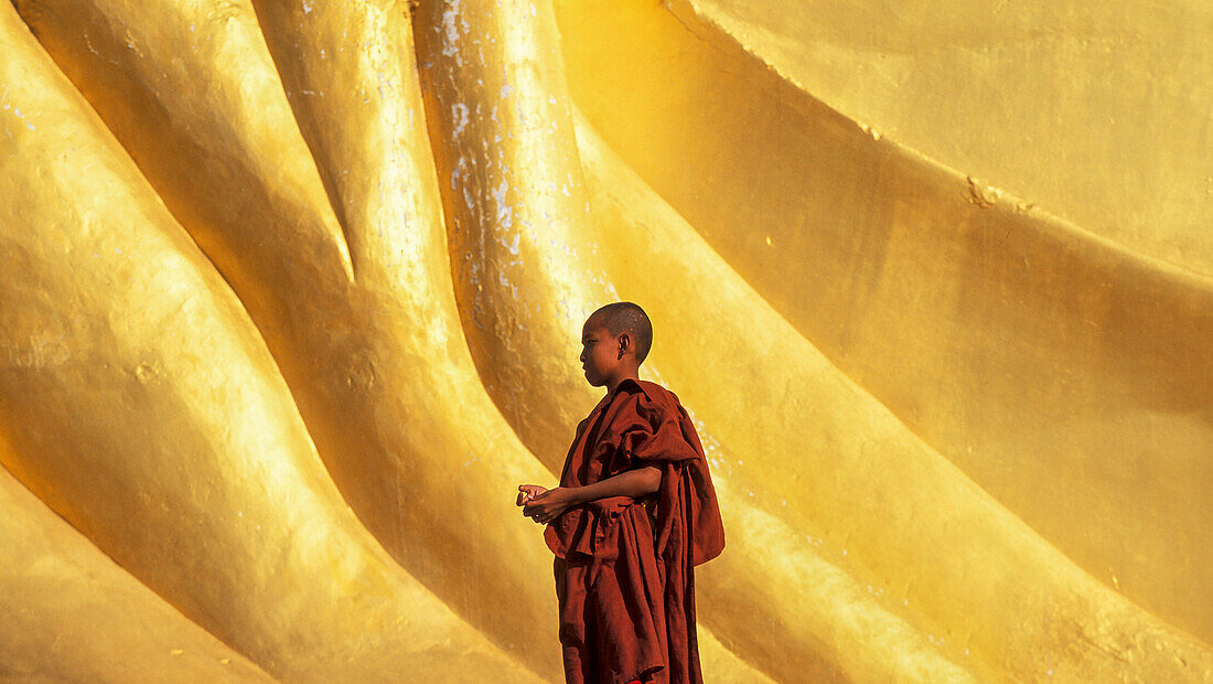 Myanmar, Monyma, Mandalay Division, Novice monk praying under giant statue of reclining Buddha in Lay Kyune Sakkyar temple