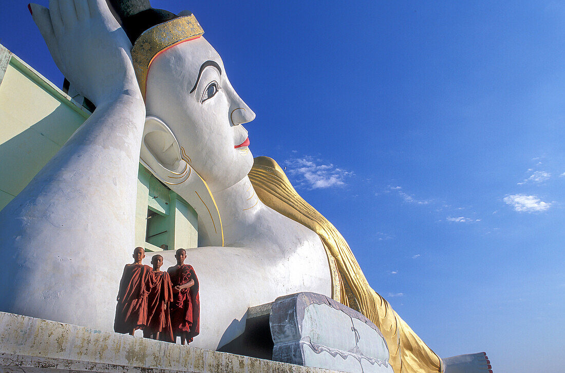 Myanmar, Monyma, Mandalay District, Novice monks standing under giant statue of reclining Buddha in Lay Kyune Sakkyar temple