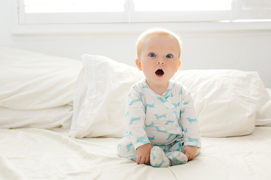 Babyjunge mit offenem Mund auf dem Bett sitzend