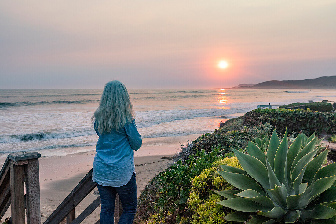 USA, Kalifornien, Cayucos, Frau beobachtet Sonnenuntergang von einer Klippe über dem Strand