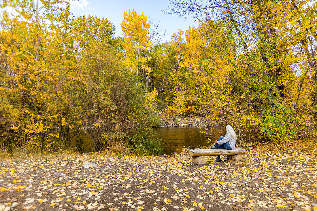 USA, Idaho, Hailey, Rückansicht einer älteren Frau, die sich im Herbst auf einer Bank am Fluss entspannt