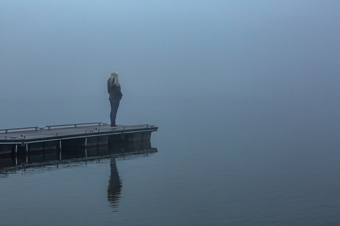 Ältere Frau steht am Morgen auf einem Steg im Nebel