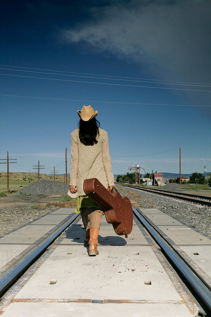 Woman with guitar walking down railroad tracks, rear view
