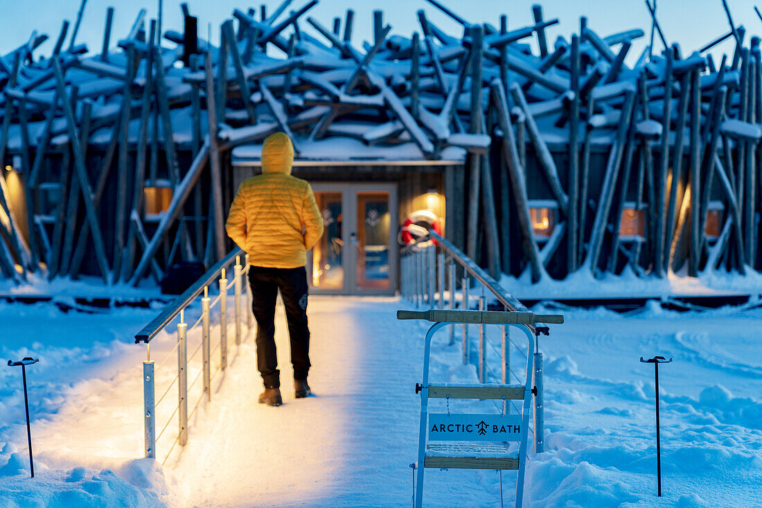 Person, die in der Abenddämmerung auf dem gefrorenen Steg steht, der das schwimmende Arctic Bath Hotel mit dem Ufer verbindet, Harads, Lappland, Schweden, Skandinavien, Europa