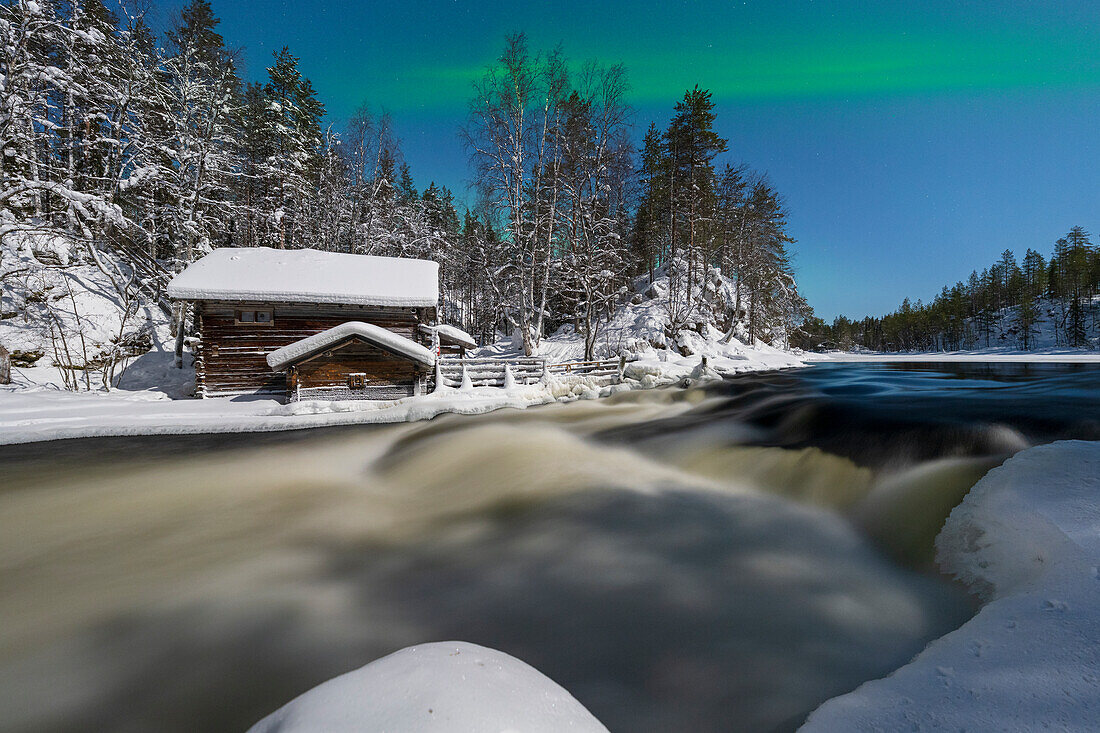 Einsame Hütte im verschneiten Wald mit Blick auf die Stromschnellen des zugefrorenen Flusses Myllikoski unter Nordlichtern, Ruka Kuusamo, Lappland, Finnland, Europa