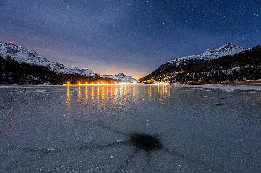 Holes in the cracked ice on Lake Champfer with Silvaplana village on background at night, Engadine, Graubunden, Switzerland, Europe