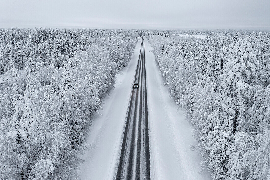 Blick von oben auf ein Auto, das auf einer glatten, leeren Straße im verschneiten Wald fährt, Lappland, Finnland, Europa