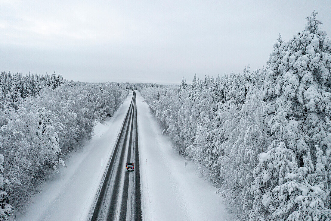 Blick von oben auf ein Auto, das auf einer vereisten Straße im verschneiten Wald fährt, Lappland, Finnland, Europa