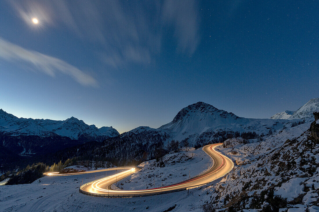 Lichter von Autospuren auf kurvenreicher, verschneiter Straße bei Nacht, Berninapass, Val Poschiavo, Kanton Graubünden, Schweiz, Europa