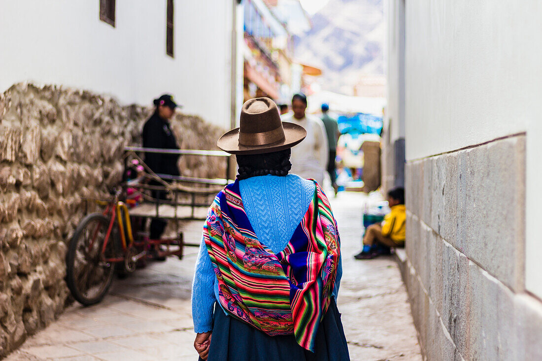 Frau geht in enger Straße mit traditioneller bunter peruanischer Tasche auf dem Rücken, Pisaq, Peru, Südamerika