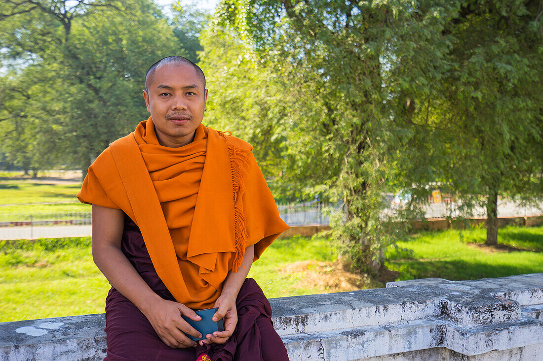 Mönch in safranfarbener Robe, der mit seinem Mobiltelefon in der Hand an der Wand sitzt, Königspalast, Mandalay, Myanmar (Burma), Asien