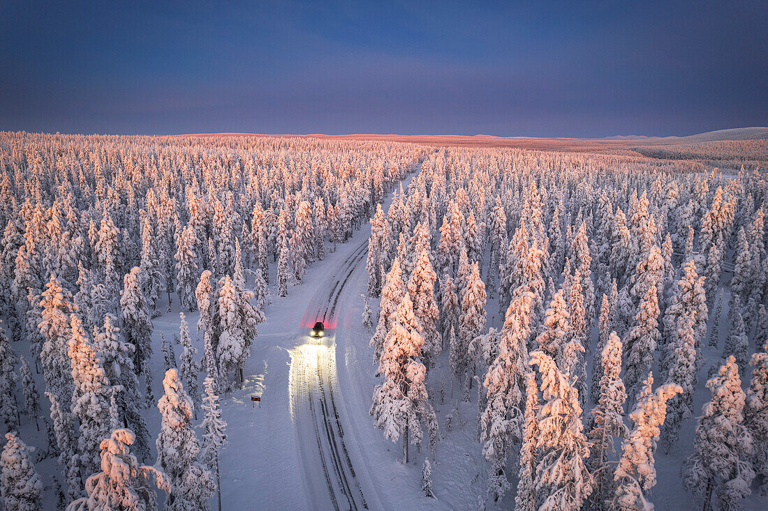 Luftaufnahme eines Autos auf vereister Straße und beleuchteten Scheinwerfern im schneebedeckten Wald, in der Morgendämmerung, Akaslompolo, Kolari, Pallas-Yllastunturi-Nationalpark, Region Lappland, Finnland, Europa
