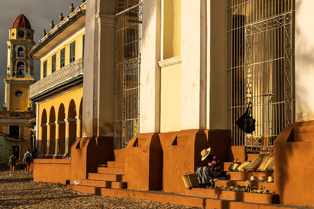 Evening sunlight bathes buildings of main square and souvenir seller on Cathedral steps, Trinidad, Cuba, West Indies, Caribbean, Central America
