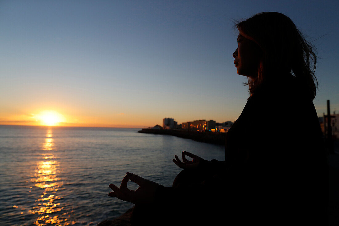 Frau praktiziert Yoga-Meditation am Meer bei Sonnenuntergang als Konzept für Stille und Entspannung, Cadiz, Andalusien, Spanien, Europa