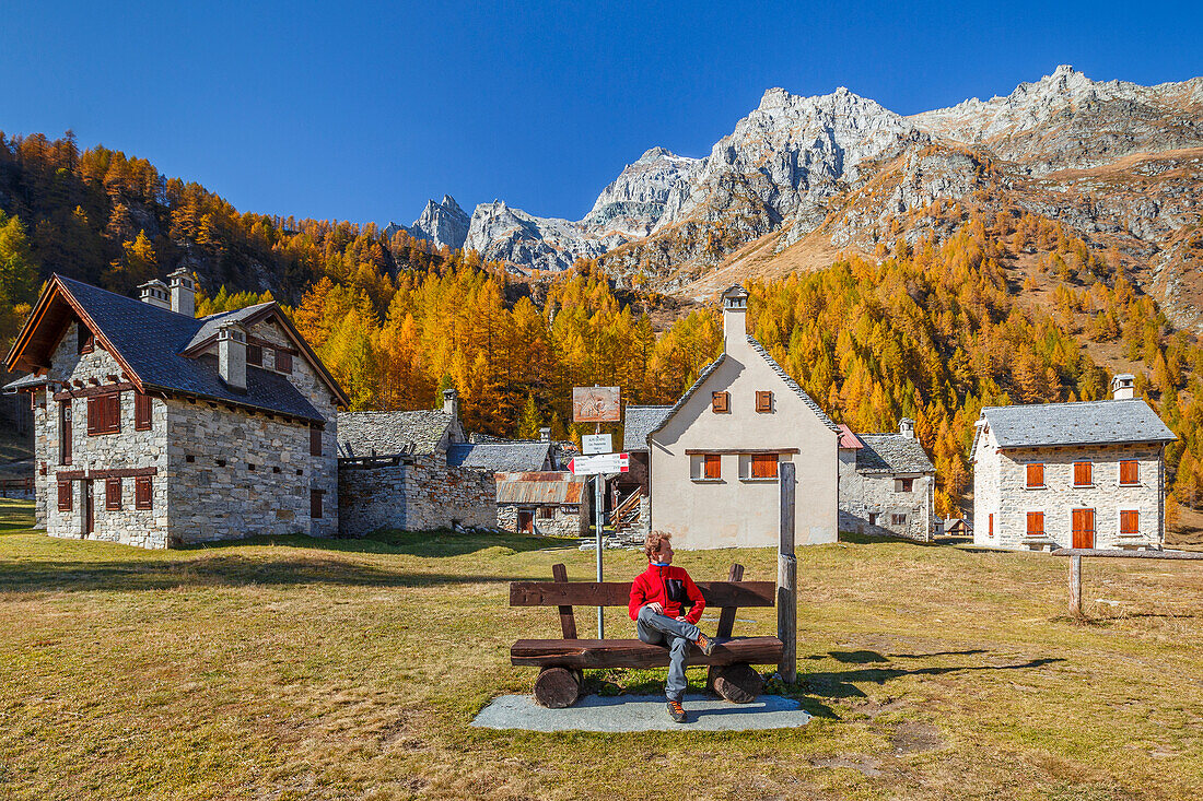 Wanderer entspannt sich auf einer Bank des Dorfes Pedemonte in der Herbstzeit, Naturpark Alpe Devero, Baceno, Alpe Veglia und Alpe Devero, Provinz Verbano-Cusio-Ossola, Piemont, Italien, Europa (MR)