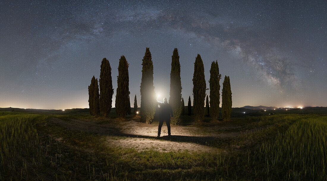 eine Person beobachtet die Milchstraßenbögen, in der Nähe der ikonischen Zypressen von San Quirico d'Orcia, Provinz Siena, Toskana, Italien, Europa