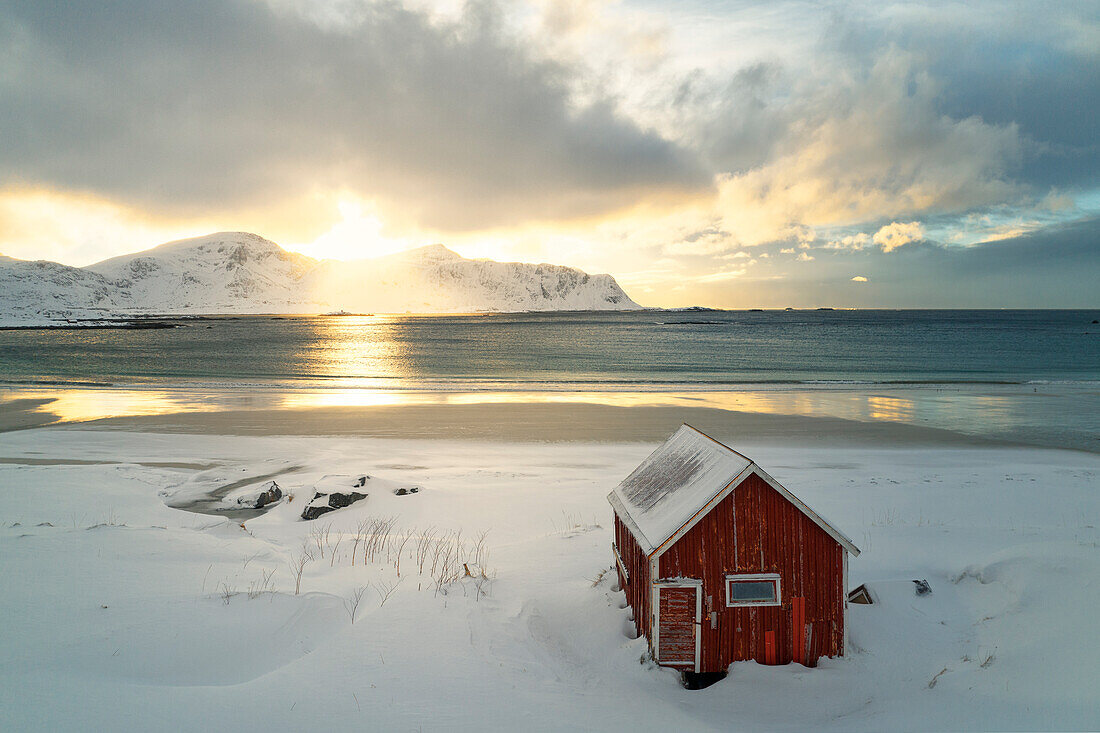 Luftaufnahme einer klassischen Rorbuer im Schnee am Strand von Ramberg während eines winterlichen Sonnenuntergangs, Ramberg, Lofoten, Norwegen, Europa