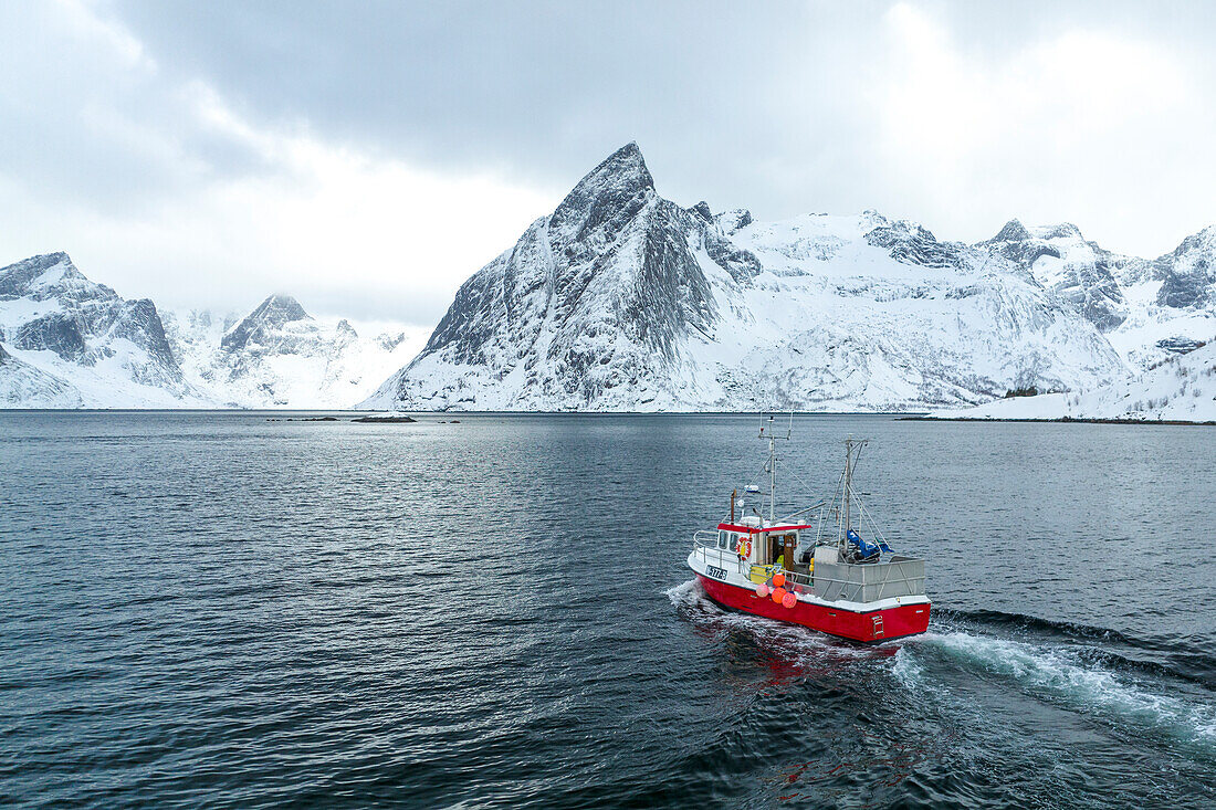 Ein Fischerboot segelt in der Bucht von Reine mit dem schneebedeckten Gipfel des Olstind, Lofoten-Inseln, Norwegen, Europa