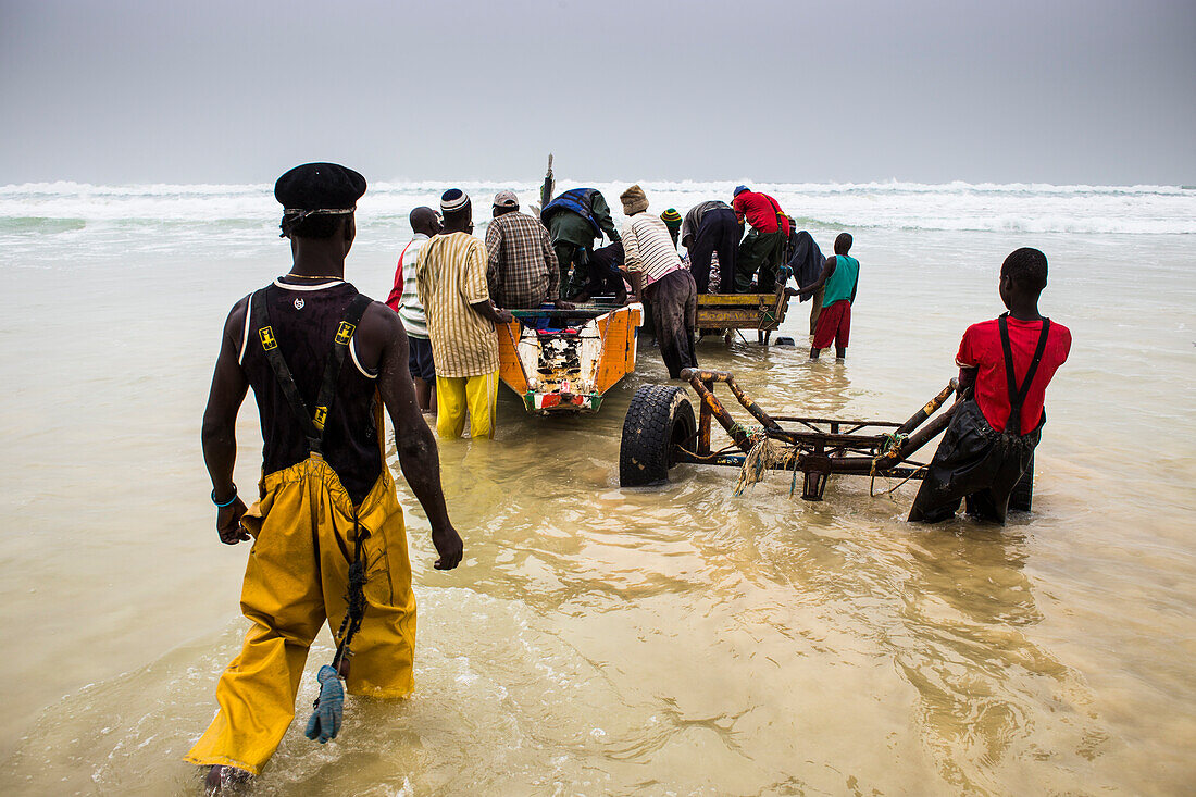 Männer entladen ein Fischkanu auf dem nachmittäglichen Fischmarkt am Strand von Mboro Plage, wenn die Kanus beladen ankommen