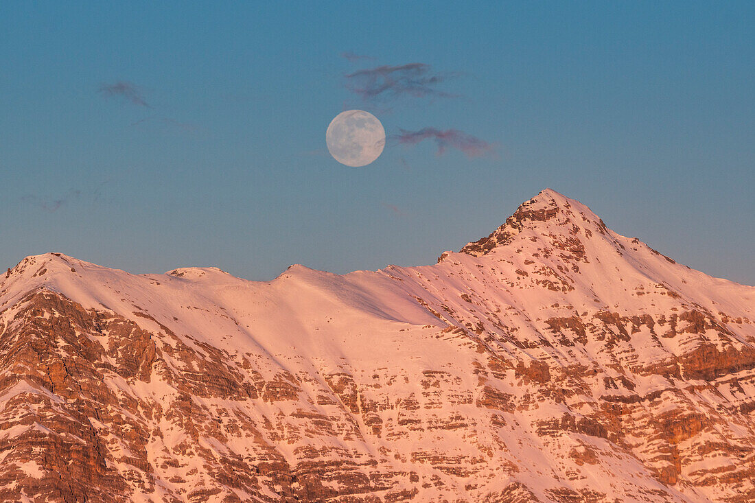 A peak illuminated from the red light of sunset with full moon just over in the sky during winter. Rocca mount, Valdidentro, Lombardy, ALps, Italy, Europe.