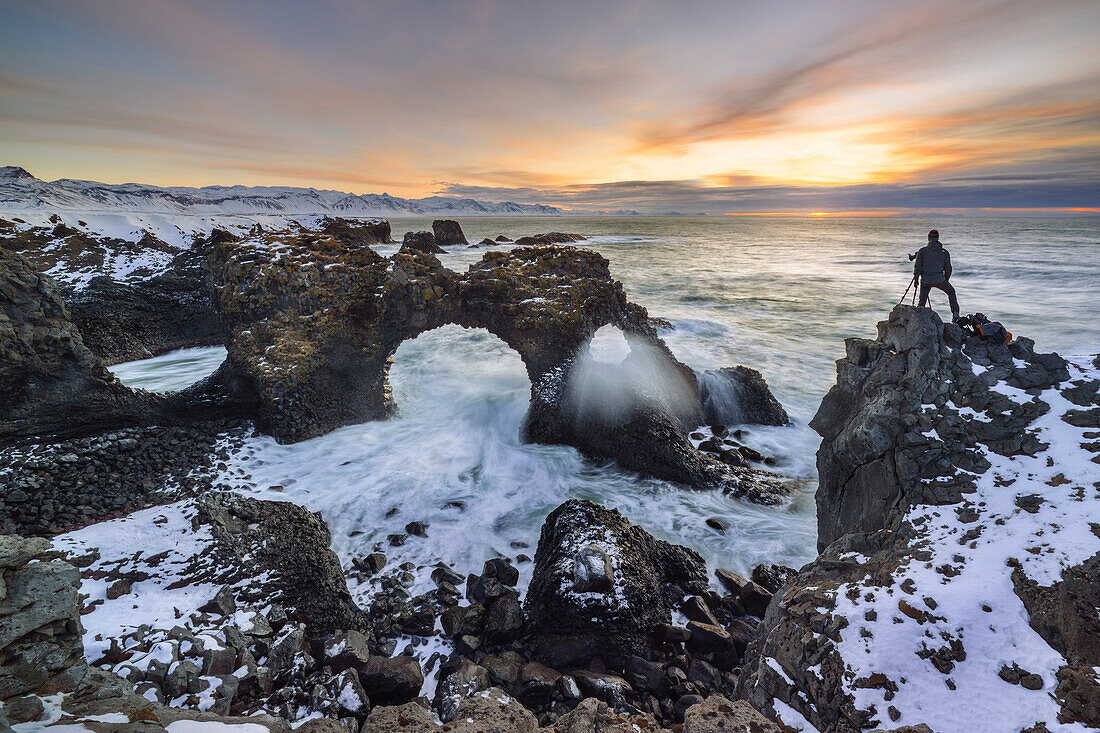 Ein Fotograf am Gatklettur-Bogen während des Sonnenaufgangs im Winter, Arnarstapi, Hellnar, Snæfellsnes-Halbinsel, Island, Nordeuropa (MR)
