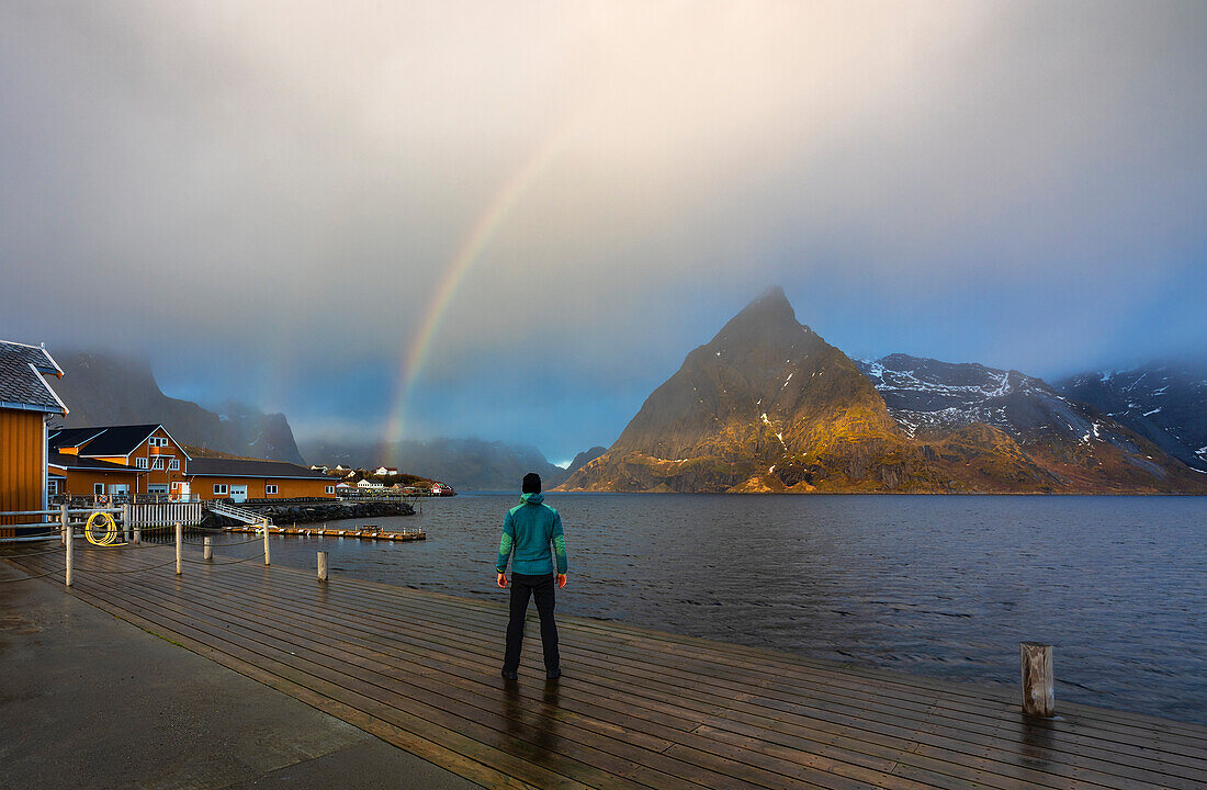 Ein Mann über einem Steg vor Sakrisoy und Olstind mit Regenbogen, Moskenes, Moskenesoya, Nordland, Lofoten, Norwegen, Nordeuropa