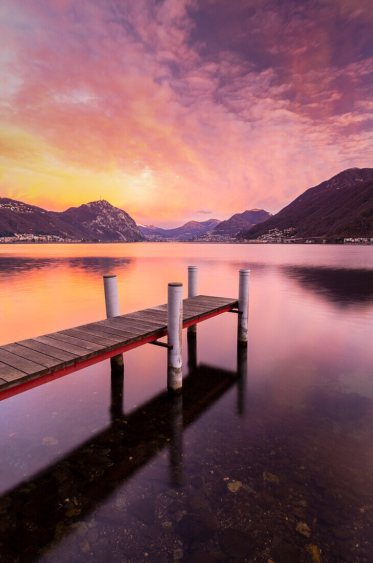 Sonnenaufgang Explosion der Farben vor Lugano und Campione d'Italia von einem Pier auf dem Ceresio-See, Riva San Vitale, Kanton Tessin, Schweiz.
