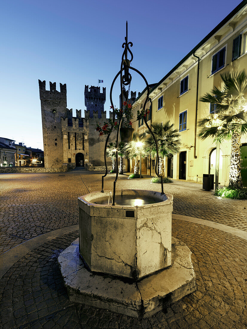 Castello-Platz in Sirmione, mit beleuchtetem Scaliger-Schloss und mittelalterlichem Brunnen; Gardasee, Sirmione, Provinz Brescia, Lombardei, Italien, Europa