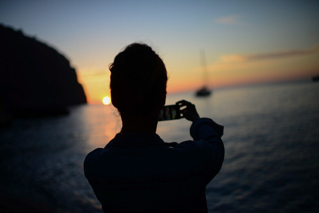 Silhouette einer jungen Frau, die den Sonnenuntergang und ein Segelboot am Strand fotografiert