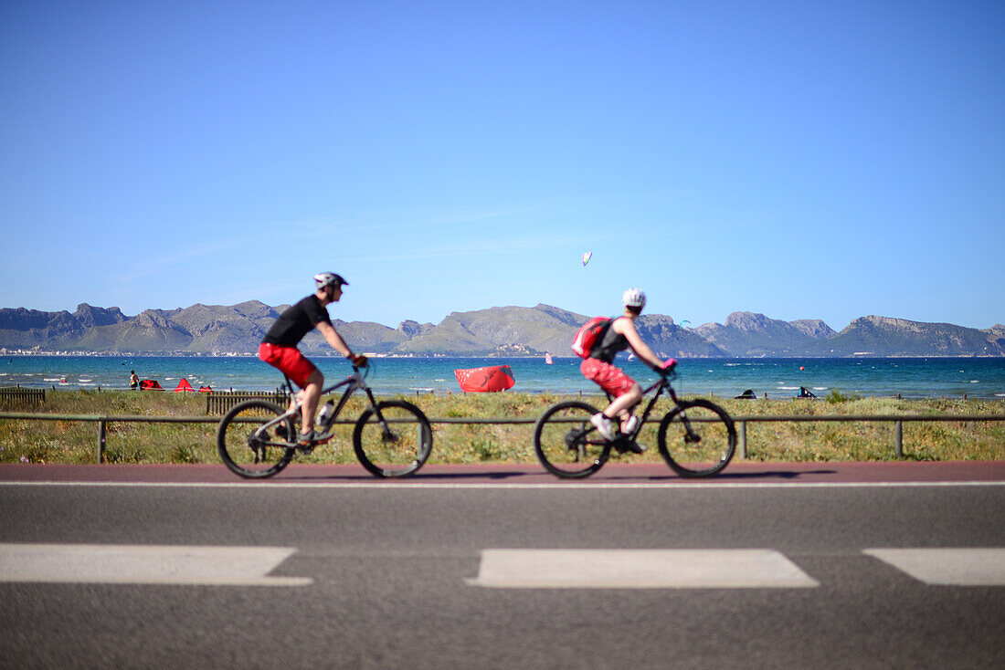 Ein Paar Radfahrer und Kitesurfer am Strand von Port de Pollenca, Mallorca, Spanien
