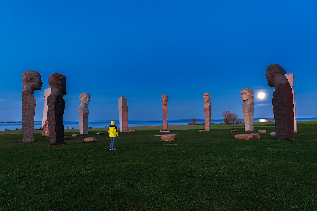 Man with yellow jacket admiring the full moon rising behind the stone statues of Dodekalitten at dusk, Lolland island, Zealand, Denmark, Europe