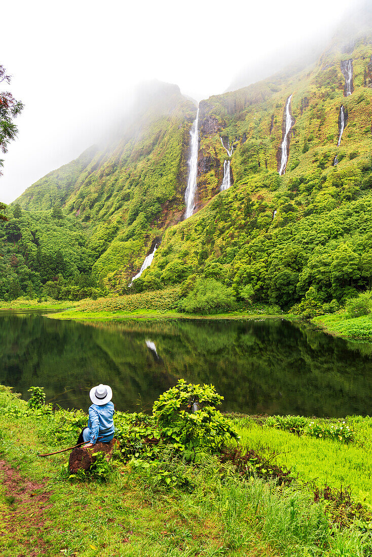 Rückansicht einer Frau mit blauem Tuch und weißem Hut, die sitzt und den Wasserfall Ribeira do Ferreiro inmitten von grüner und üppiger Vegetation mit Wolken bewundert, Poco Ribeira do Ferreiro (Alagoinha), Gemeinde Lajes das Flores, Insel Flores (Ilha das Flores), Azoren-Archipel, Portugal, Europa