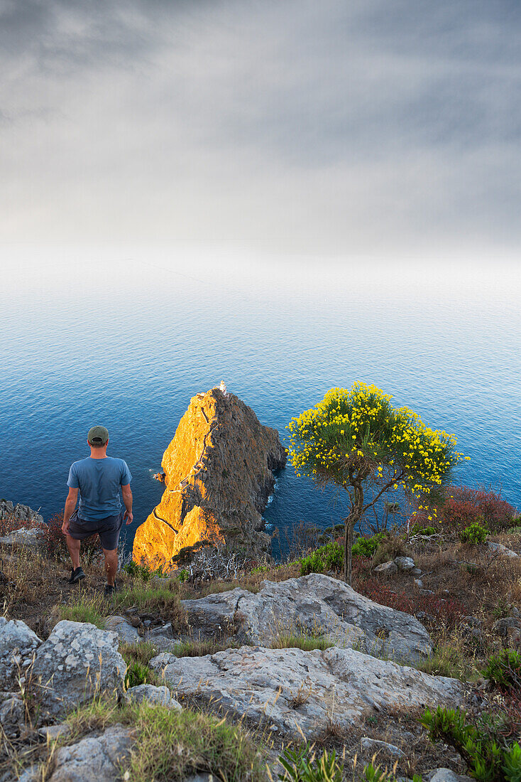 Blick von oben auf einen Mann, der auf dem Gipfel des Monte della Guardia steht und den Leuchtturm Punta della Guardia bei Sonnenaufgang überblickt, Insel Ponza, Archipel Pontino, Provinz Latina, Latium, Italien