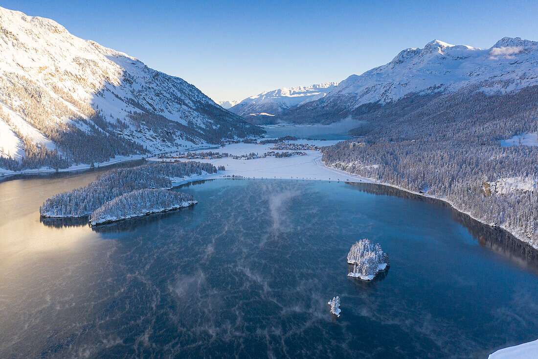 Luftaufnahme von verschneiten Wäldern und Sils Maria am Ufer des zugefrorenen Silsersees, Graubünden, Engadin, Schweiz