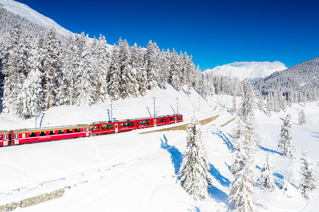 Wintersonne über dem Bernina Express Zug, der durch die verschneite Landschaft fährt, Chapella, Kanton Graubünden, Engadin, Schweiz